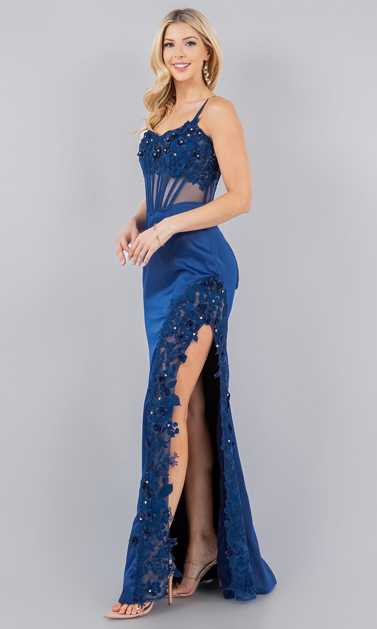 Woman standing and smiling in the Cinderella Couture 8085J Long Corset Prom Dress, a sleeveless blue gown with lace detailing, beaded embroidery, high slit, and strappy heels on a plain background.